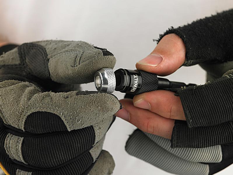 A close-up of gloved hands holding a DEWALT drill bit holder, showing the textured silver collar and black body with the DEWALT logo.