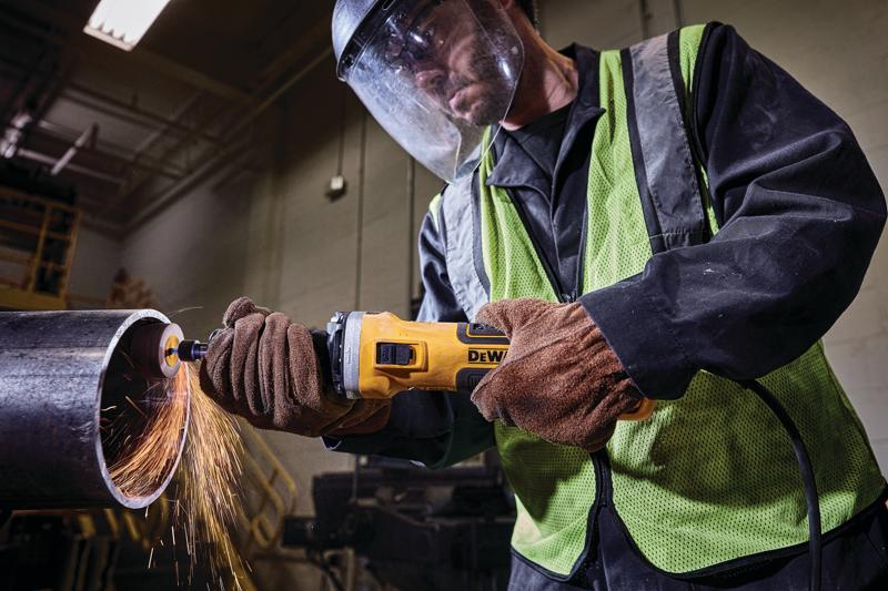 A person wearing protective gear and gloves uses a DEWALT angle grinder to cut through a large metal pipe, with sparks flying from the cutting area.